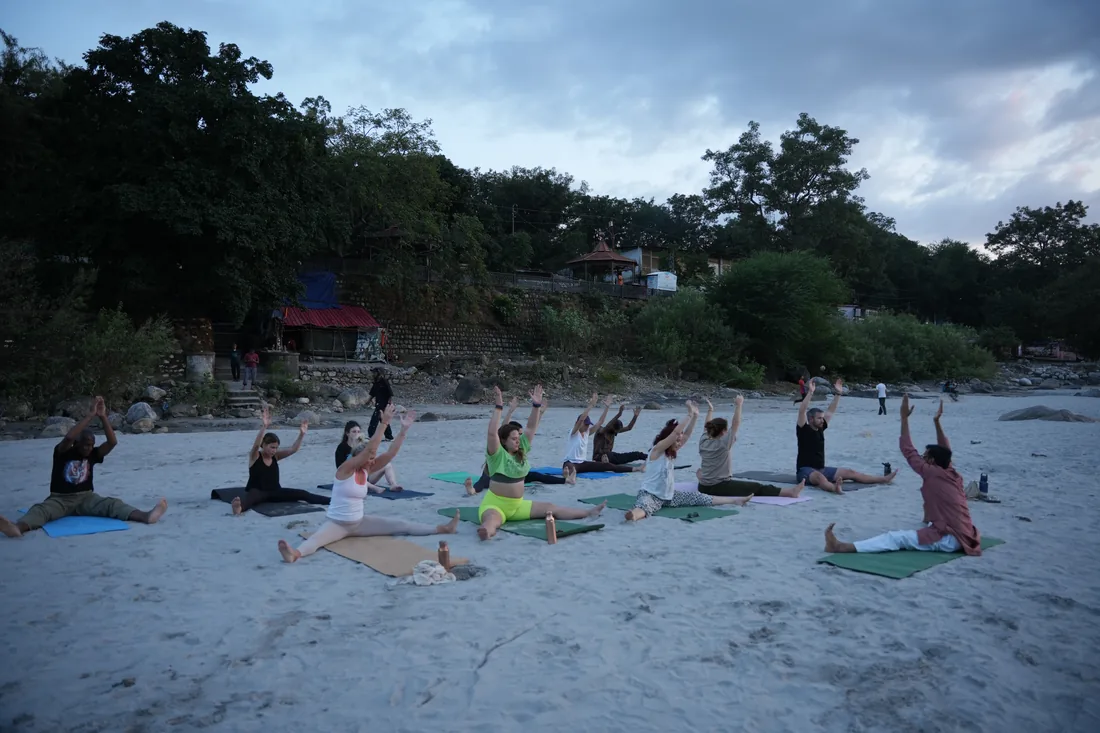 Sri Yoga Ashram group class raising arms in seated pose on Rishikesh river sand